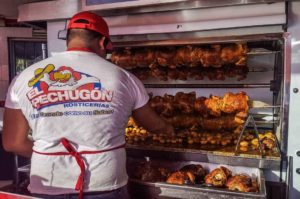 An employee in an El Pechugon shirt in front of a spit with whole roast chickens and roasted potatoes