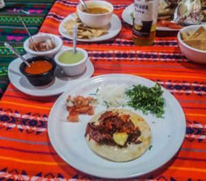 One al pastor taco on a plate with small portions of cilanto, onions and pico de gallo on the side. All on a brightly coloured Mexican blanket