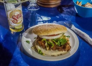 Plate with a chicken burger with lettuce and onion on a blue table cloth with a Sol beer
