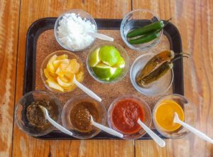 A tray with taco toppings in little bowls. 4 kinds of salsa, 2 kinds of peppers, limes, lemons and onions