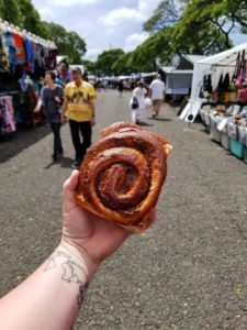 Hand with a map tattoo on the inner wrist holding a giant cinnamon roll in front of booths