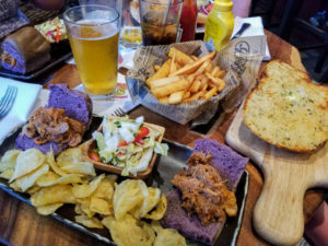 Pulled pork sliders with a side of coleslaw and a side of fresh potato chips beside a loaf of garlic bread, an order of fries and a beer