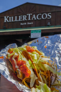 2 tacos in a foil wrap being held up in front of the Killer Tacos sign