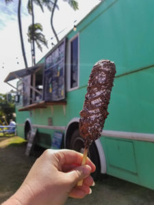 Hand holding a waffle on a stick in front of a teal food truck