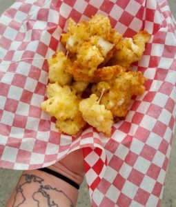 Deep fried cheese chunks on a red & white checkered paper being held by a hand with a tattoo of the world on the inner wrist
