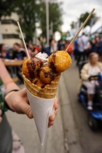 Donut holes on skewers in a waffle cone with whipped cream. Cone is held by a hand with a festival in the background