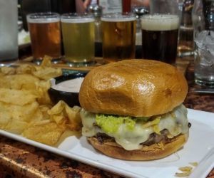 In the foreground a beef burger topped with extra pepper jack cheese and guacamole on a closed bun with a side of fresh chips, a flight of 4 beers in the background at Black Iron Grill in Miles City, Montana