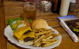  Open faced double cheeseburger with a side of fries. Lettuce, tomato, onion and pickle are on the side. A mason jar of beer sits behind the plate at Powder River Stockman's Club in Broadus, Montana