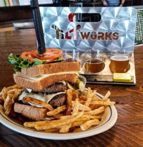 2 cheeseburger patties between 2 grilled cheese sandwiches, with a steak knife in the top holding lettuce and 2 tomatoes. A metal flight rack with the 'Tiltwurks' logo at Tiltwurks in Miles City, Montana