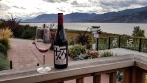 A bottle of Bench 1775 wine and a glass of red on a railing with Okanagan Lake in the background