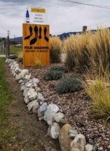 A line of shrubs surrounded by wood chips and a rock wall, in front of the Four Shadows Winery entry sign on Naramata Bench