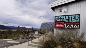 The Monster Vineyards tasting room sign, welcoming skeptics and believers. With a view of Okanagan Lake in the background