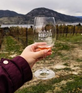 A hand holding a wine glass with the Monster Vineyards logo on it, with vineyards and Okanagan Lake in the background