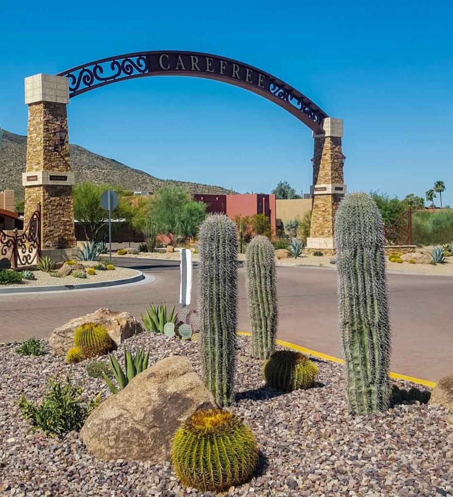 A group of cacti, including 3 tall ones, stand in front of a Carefree town sign arched above a roadway