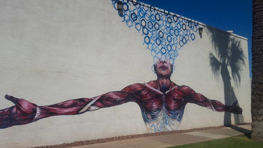 Street art on a white wall. A man with no skin and only muscles showing, with arms outstretched an the top half of his head giving way to an explosion of hexagons in shades of blue