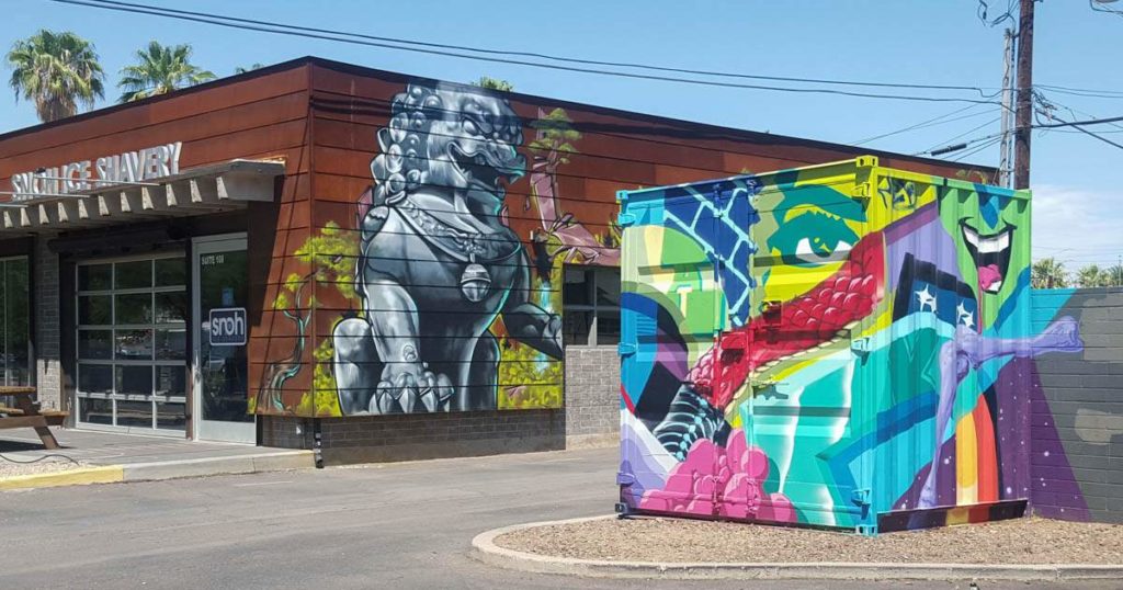 An ornate grey stone lion painted on the side of a brown building with a brightly coloured storage unit in the foreground