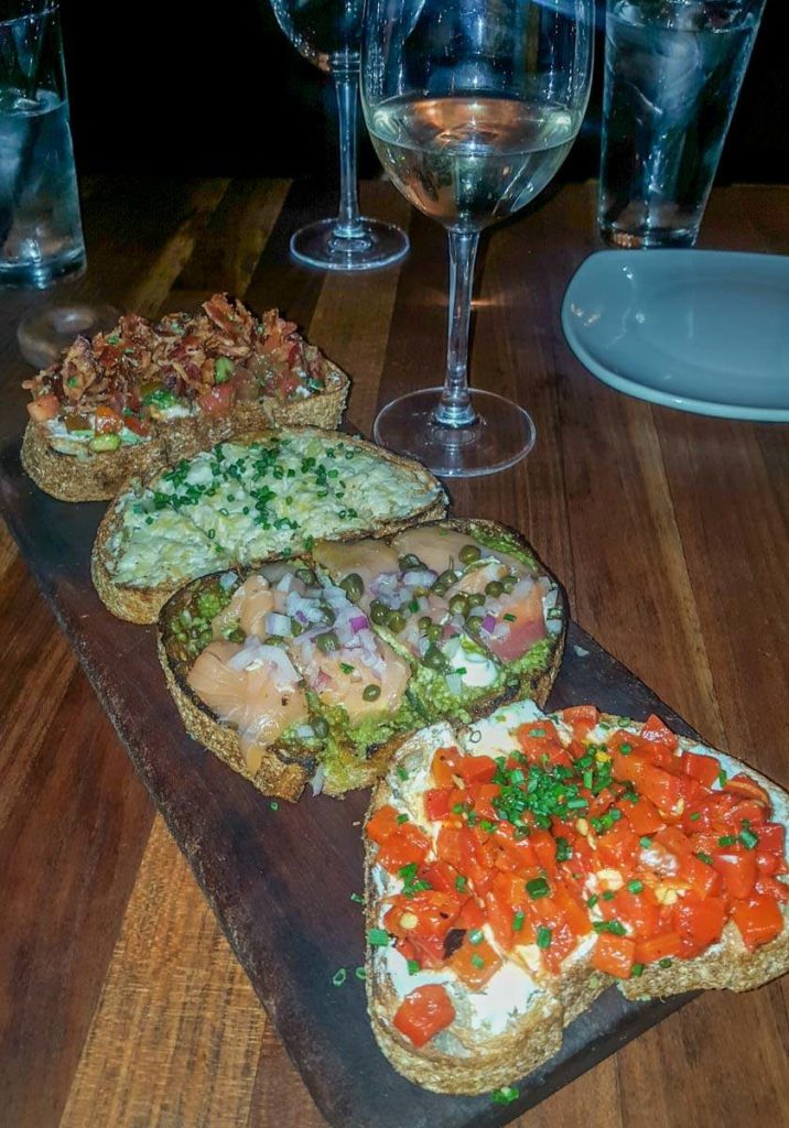 A tray with 4 pieces of toast, each topped with different ingredients. Including bruschetta, bacon, avocado, salmon and artichoke dip with 2 glasses of wine set behind it. 