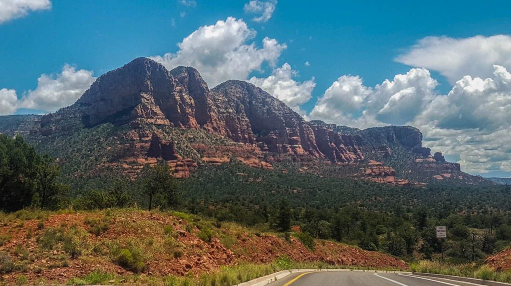 A red rock mountain looms over a piece of highway with a blue sky and puffy white clouds framing it. 
