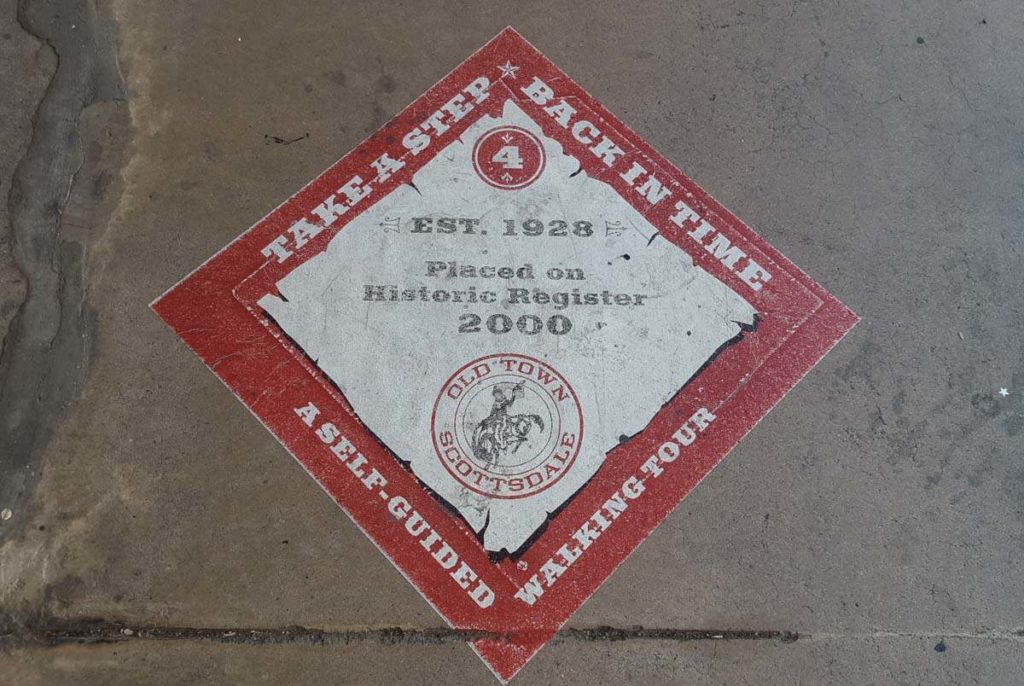A diamond sign painted on a sidewalk with the words 'Take a step back in time, a self-guided walking tour' in a red band around the outside and '4- Est 1928, Placed on Historic Register 2000' in the white center