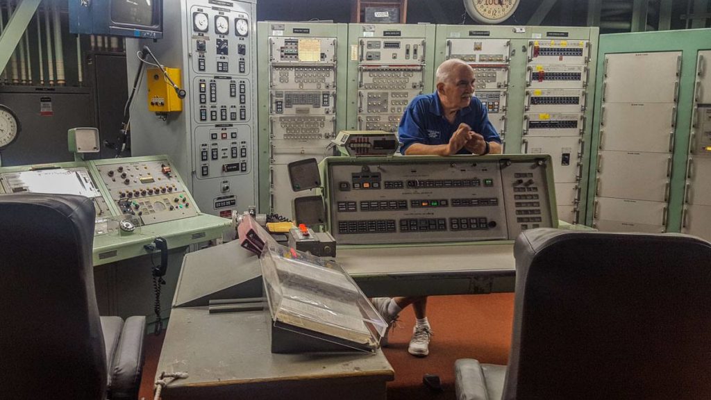 A man in a blue shirt standing in front of a wall of control panels. He is leaning on another desk top set of controls. 