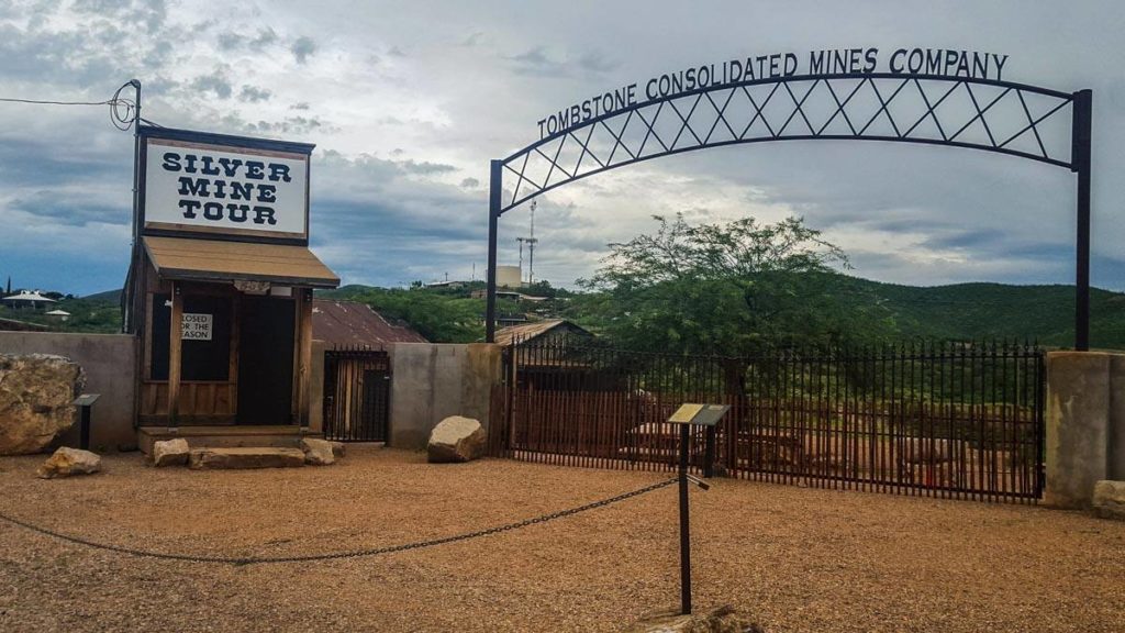 A ticket booth labelled 'Silver Mine Tour' stands beside a metal archway with 'Tombstone Consolidated Mines Company' written across it. The gate under the arch is closed and both are behind a chain gate