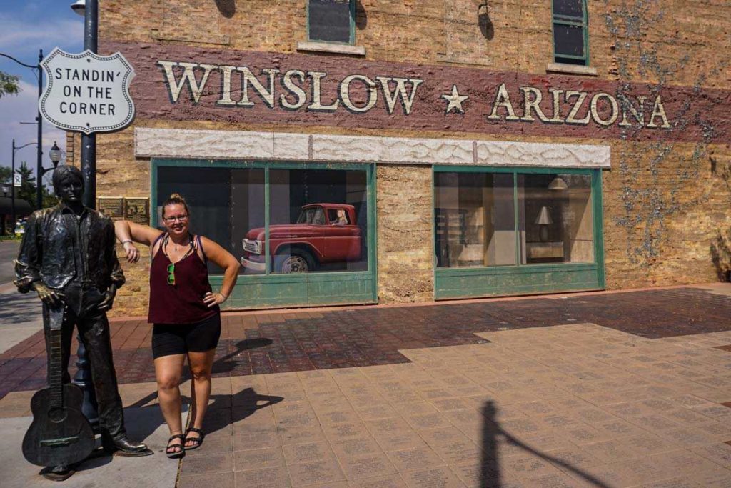 A woman with her elbow on the shoulder of the metal sculpture named 'Easy', underneath a sign that says 'standin' on the corner'. A painted mural or a red trunk and a Winslow, Arizona sign are behind her.