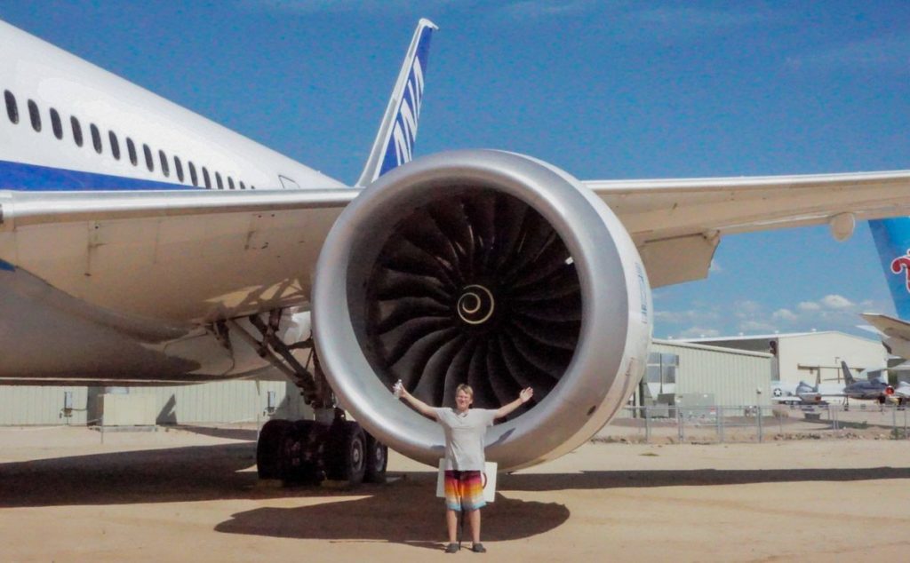 A boy standing in front of a plane engine with his arms outstretched to show how big it is